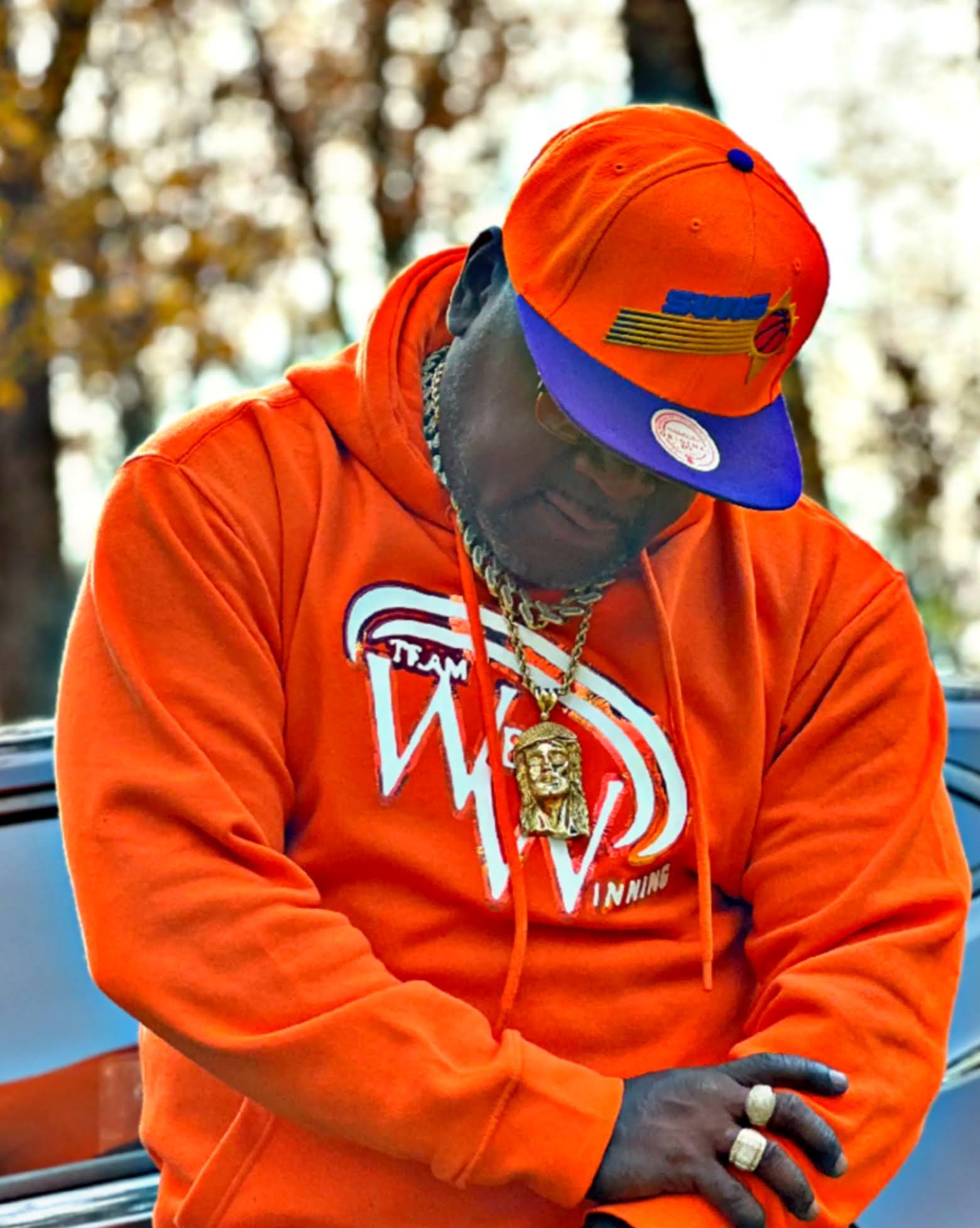 Person wearing orange Team We Winning hoodie and cap, standing outdoors near a car